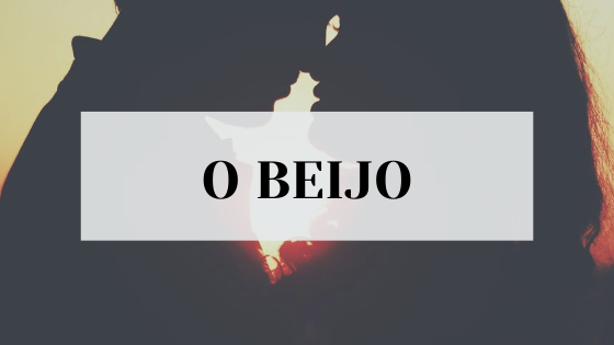 #pracegover audiodescrição resumida: foto de um casal na contraluz. Seus rostos estão próximos, seus narizes se tocam. No centro, retângulo branco com o escrito "O Beijo".