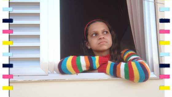 Foto de Natália com 11 anos no parapeito de uma janela, olhando para cima. Natália está usando uma blusa de frio listrada, colorida.